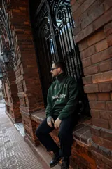 Buzo verde oscuro de algodón con cierre medio frontal, cuello alto y corte oversize. Estampado frontal con la frase "Against All" en color crema y detalles bordados.