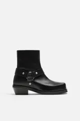Bota negra de cuero con caña corta, horma cuadrada, suela a tono con ligero tacón y arnés decorativo a la altura del tobillo con detalles metálicos.