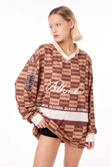Camiseta oversize marrón con estampado de logos y texto "Blkmba" en toda la prenda. Tiene cuello en V con cuello tejido, escudo Mamba soccer en la manga y logo blkmba en el centro.