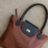 Cartera tote de cuero vacuno color marrón con solapa negra y cierre metálico. Tiene doble asa de mano negra, bolsillo interno con cierre y cierres YKK metálicos.