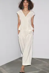 Pantalón culotte color crudo, de tiro medio, confeccionado en lino. Presenta bolsillos laterales, pinzas delanteras, pierna ancha y cierre frontal con cremallera, botón y ganchos metálicos.