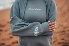 Hoodie gris con capucha y cordones ajustables, de corte holgado y manga larga. Presenta un bordado de flores blancas en la espalda y un pequeño bordado en la manga.