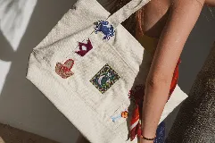 Bolso tote de lona color crudo con asas de hombro. Decorado con parches bordados de motivos playeros y pañuelo rojo estampado atado a una de las asas.