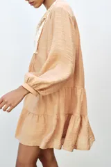Vestido corto color beige con cuello redondo, cordón con borlas y manga larga. Presenta bordados combinados a contraste en la pechera, cierre frontal con botones ocultos, forro interior y bajo con volantes.