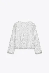 Chaqueta corta blanca con lentejuelas plateadas, cuello redondo, manga larga y forro interior.