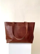 Cartera tipo tote de cuero graneado color marrón, con manijas de 55 cm de largo. Tiene un bolsillo grande en el frente con broche de imán y un bolsillo con cierre metálico en la parte posterior. El cerramiento principal es mediante un cierre metálico. En su interior, está forrada y cuenta con tres bolsillos, uno con cierre, otro plaqué y un tercero grande tipo sobre con solapa y botón.