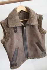 Chaleco negro de cuero con cuello y borde inferior de piel sintética negra. Cierre frontal con cremallera color cobre y detalles en cuero color tostado.