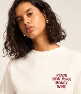 Remera blanca de algodón con corte boxy, cuello redondo y mangas cortas con dobladillo. Presenta un bordado en el pecho con la frase "PEACE NEW YORK MONEY WINE" en color borgoña.