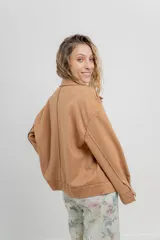 Chaqueta color beige de corte recto, estilo camisa, confeccionada en gamuza con elastano. Presenta cuello camisero, cierre frontal con botones metálicos, bolsillos grandes en el frente y charreteras en los hombros.