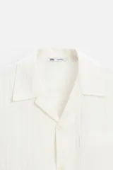 Camisa color blanco con textura de rayas verticales, corte relaxed fit, cuello bowling, manga corta, bolsillo de parche en el pecho y cierre frontal con botones.