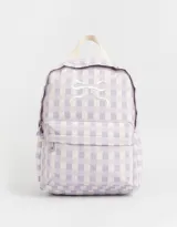 Mochila de tela con estampado a cuadros lila y blanco, con bolsillo frontal con cierre y moño bordado. Tiene correas regulables, manija superior y doble cierre superior.
