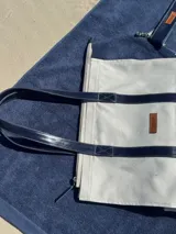 Bolso de lona color blanco con asas de cuero azul marino y etiqueta de cuero con el logo de la marca.