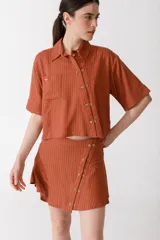 Camisa corta color ladrillo con rayas verticales blancas, de manga corta, con cuello clásico y doble cartera diagonal con botones de madera. Presenta un aplique de crochet hecho a mano en el bolsillo.