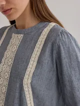Camisa celeste de algodón con detalle de encaje blanco en el frente. Tiene mangas largas abullonadas con puños anchos y cuello redondo.