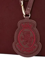 Cartera tipo tote bag de color borgoña, con acabado liso y detalles en cuero sintético del mismo tono. Presenta dos asas superiores y una correa ajustable extraíble. Incluye un colgante decorativo con el escudo de Hilfiger bordado y un dije metálico con el monograma TH.