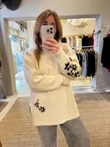 Sweater de punto color beige con cuello alto, mangas largas y corte holgado. Presenta tajos laterales y bordados florales en puños y cadera.