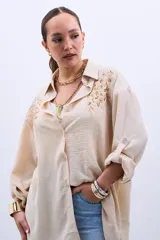 Camisa color beige con bordado de lentejuelas doradas en los hombros, corte clásico, cuello camisero y mangas largas con puños abotonados.