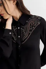 Camisa negra de manga larga, estilo texana, con cuello camisero y botones metálicos tipo snap. Presenta un bordado de tachas termo fijadas en forma de pico en la zona del pecho y hombros.