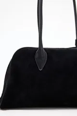 Cartera tipo bowling negra, confeccionada en material que simula gamuza o ante, con dos asas superiores de cuero liso negro. Presenta una forma alargada y redondeada en la base.