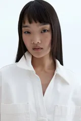 Vestido corto blanco de denim rígido, corte recto y holgado, con cuello camisero, escote en V, mangas cortas y bolsillos de parche en el pecho.