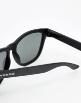 Gafas de sol unisex Hawkers, con montura cuadrada negra con textura y lentes espejados azules.
