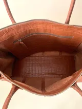 Bolso tote grande de cuero color bordó, con estructura rígida y doble asa de hombro. Presenta una tira con tachas y hebillas en cada extremo, y lazos en los costados.