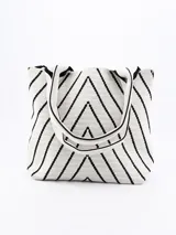 Bolso tipo tote de tejido de punto color crudo con diseño de líneas diagonales negras formando figuras geométricas. Tiene doble asa corta de hombro.