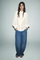 Camisa blanca semitransparente con cuello subido, manga larga con puños, pliegues y volantes combinados. Cierre frontal con botones.