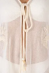 Salida de playa corta tipo poncho en color blanco marfil, confeccionada en algodón ligero y traslúcido. Presenta escote en V con lazo frontal ajustable terminado en borlas y detalles bordados en tono crema.