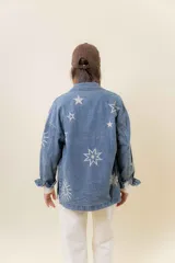 Camisa de denim de lavado medio, con calce relajado y ligeramente oversize. Presenta bordados o estampados de inspiración celestial (soles, lunas y estrellas) en color claro en la parte delantera y mangas. Tiene cuello, bolsillos delanteros con tapa y cierre frontal con botones.