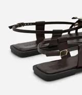 Sandalia plana color marrón con punta cuadrada, tiras finas con flor central y strass, y cierre de tira fina con hebilla cuadrada.