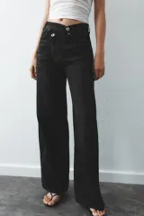 Pantalón de jean celeste de tiro alto, corte wide leg y largo hasta el tobillo. Presenta un diseño asimétrico en la cintura con cierre frontal cruzado con cremallera y botones metálicos.