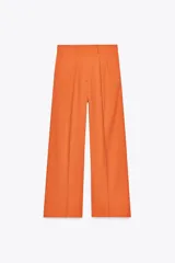 Pantalón ancho color naranja, de tiro medio y cintura con trabillas. Tiene bolsillos laterales y de vivo en espalda, pinzas en delantero, raya planchada y cierre frontal con cremallera, botón y ganchos metálicos.