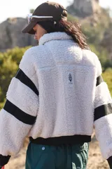 Campera de fleece de pelo alto color blanco, con cuello alto, cierre frontal completo y detalles en negro en el cierre, puños, cintura y franjas en las mangas. Incluye bolsillos frontales con solapa en color blanco liso.