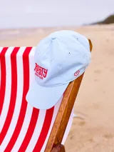 Gorra con visera color celeste con logo bordado de Rusty Beach Club en rojo.