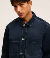 Camisa masculina oversized confeccionada en jeans azul oscuro, con cuello inglés, manga larga, puño ajustado, abotonado delantero y dos bolsillos cargo en el pecho.