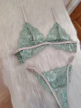 Conjunto de lencería de encaje floral color verde menta, con detalles y tiras ajustables en tono beige/tostado. Incluye un corpiño triangular sin aro y una bombacha tipo colaless o tanga.