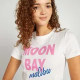 Camiseta corta blanca con estampado de letras rosas y azules con la frase "Moon Bay Malibu".