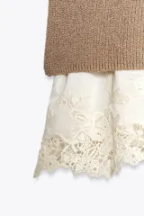 Vestido color beige, de corte recto, con cuello redondo y sin mangas. Presenta un tejido de punto en la parte superior y un volante de encaje blanco en el ruedo.