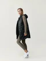 Chaleco largo acolchado negro con capucha, cierre de cremallera de doble cursor, cordones ajustables en la capucha y bolsillos con vivo. Tejido repelente al viento con aislamiento térmico y patrón holgado.