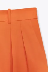 Pantalón ancho color naranja, de tiro medio y cintura con trabillas. Tiene bolsillos laterales y de vivo en espalda, pinzas en delantero, raya planchada y cierre frontal con cremallera, botón y ganchos metálicos.