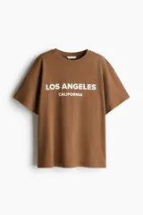 Remera color marrón con estampado blanco con la frase 'Los Ángeles California'. Corte recto y holgado, mangas cortas y cuello redondo.