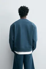 Sudadera azul petróleo de corte relajado, cuello redondo, manga larga, bolsillo tipo canguro y terminaciones en rib.