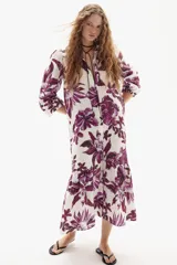 Vestido midi de lino con estampado floral grande en tonos morados sobre fondo blanco. Presenta cuello camisero, cierre frontal parcial con cordones negros y mangas tres cuartos abullonadas. La falda tiene un corte con volado en el ruedo.
