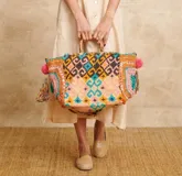 Bolso tipo tote con estampado geométrico multicolor y bordados en los laterales. Tiene asas de madera y detalles de borlas y pompones.