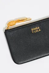 Monedero negro de cuero con cierre de cremallera y tirador de cordón amarillo. Incluye bolsillo interior y placa metálica con el logo de la marca.