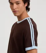 Remera de algodón de corte regular, color blanco hueso, con cuello redondo y ribete negro. Presenta un diseño de doble raya negra en las mangas y ribetes negros en el cuello y los puños de las mangas.