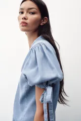 Camisa celeste de denim con cuello solapa, escote en pico, mangas abullonadas con lazos y cierre frontal con botones.