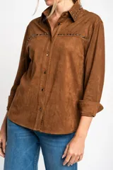 Camisa marrón de gamuza vegana con cuello clásico, cierre frontal con botones y mangas largas con puños abotonados. Presenta detalles de tachas metálicas en cuello y charreteras.