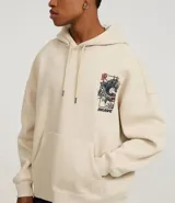 Hoodie color beige claro, corte boxy, con capucha ajustable y bolsillo canguro. Presenta un gran estampado gráfico en la espalda con un dragón oriental, texto en inglés y caracteres japoneses, y la palabra "BRAVE" en la parte superior.